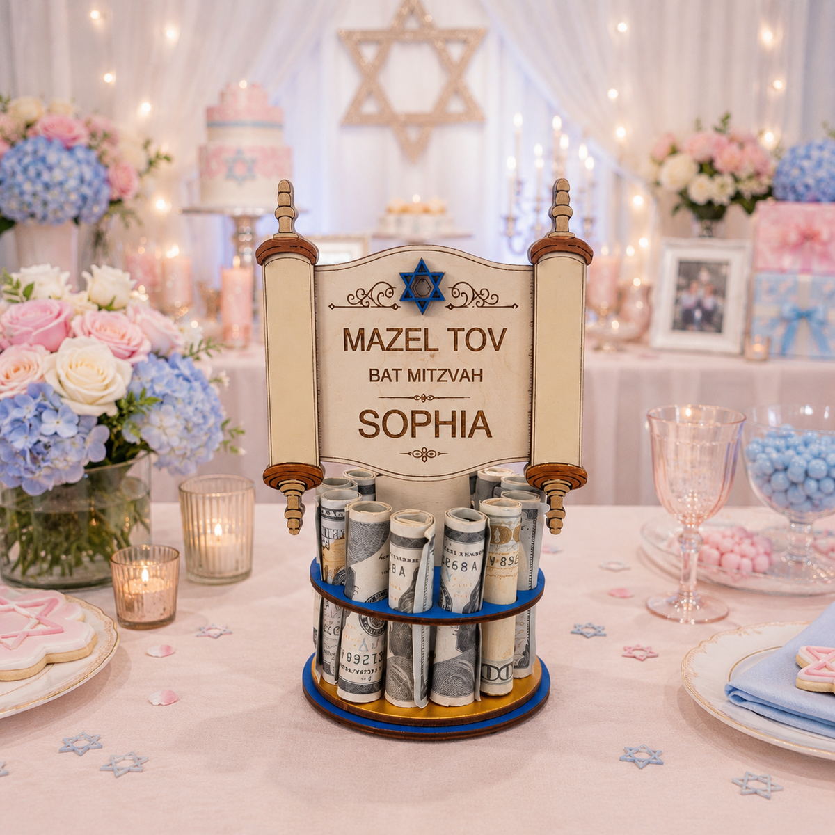 A personalized Bat Mitzvah money table featuring a scroll-shaped sign that reads 'MAZEL TOV BAT MITZVAH SOPHIA' with a blue Star of David, holding multiple rolled dollar bills on a round base. The setup is on a decorated table with pink and blue flowers, candles, and a festive background.