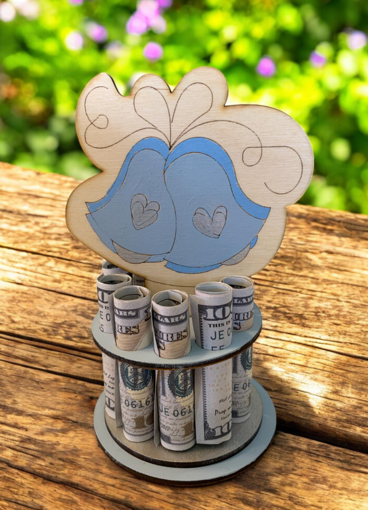 Handmade wood wedding bells money tree topped with two soft blue wedding bells and heart details, displaying rolled bills in two rows, photographed outdoors on a wood surface with greenery behind
