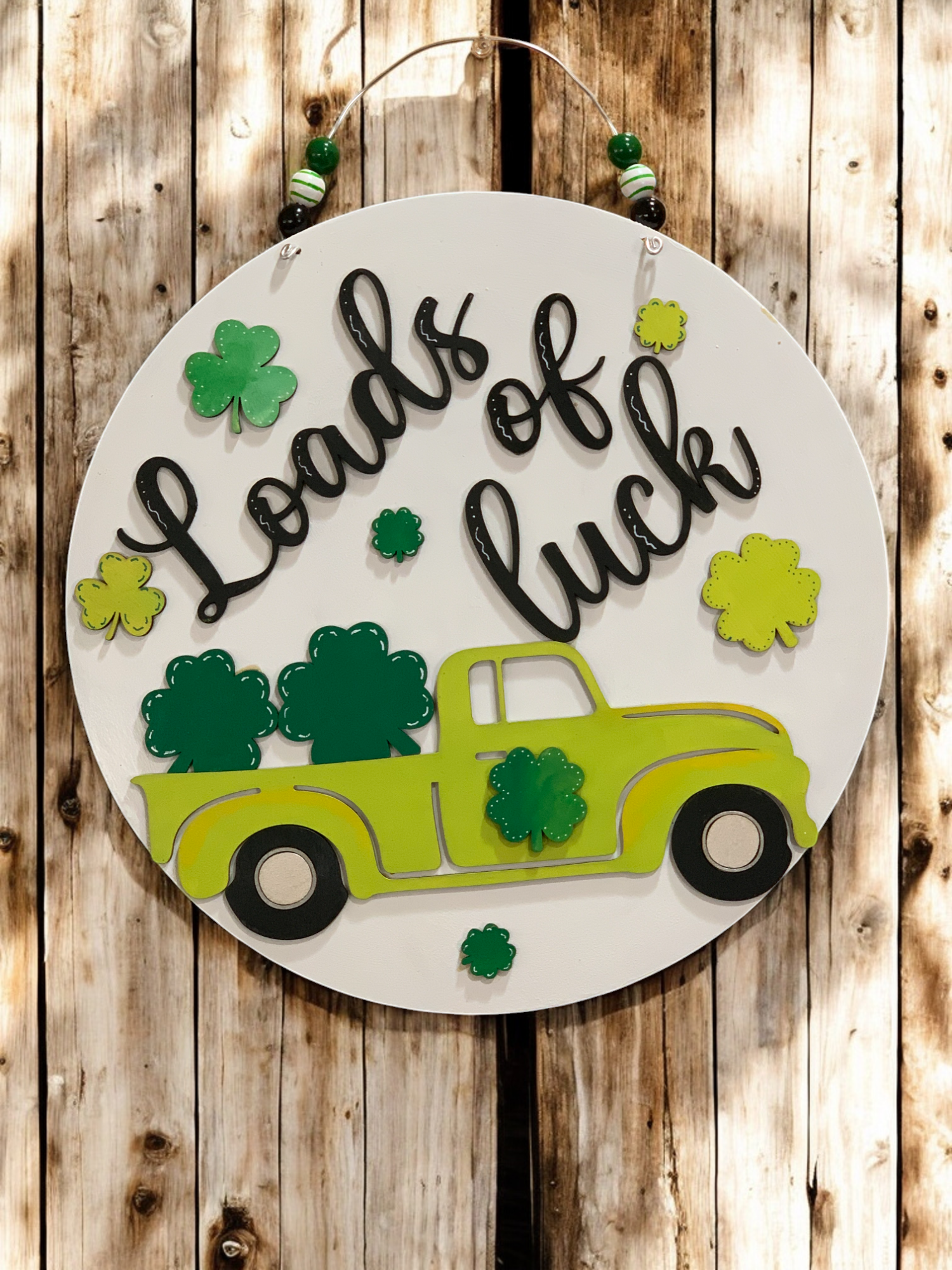 A round wooden sign with a vintage green truck design filled with shamrocks, and the text ' Loads of luck ' written in a festive font, surrounded by decorative shamrocks and a beaded wire hanger.