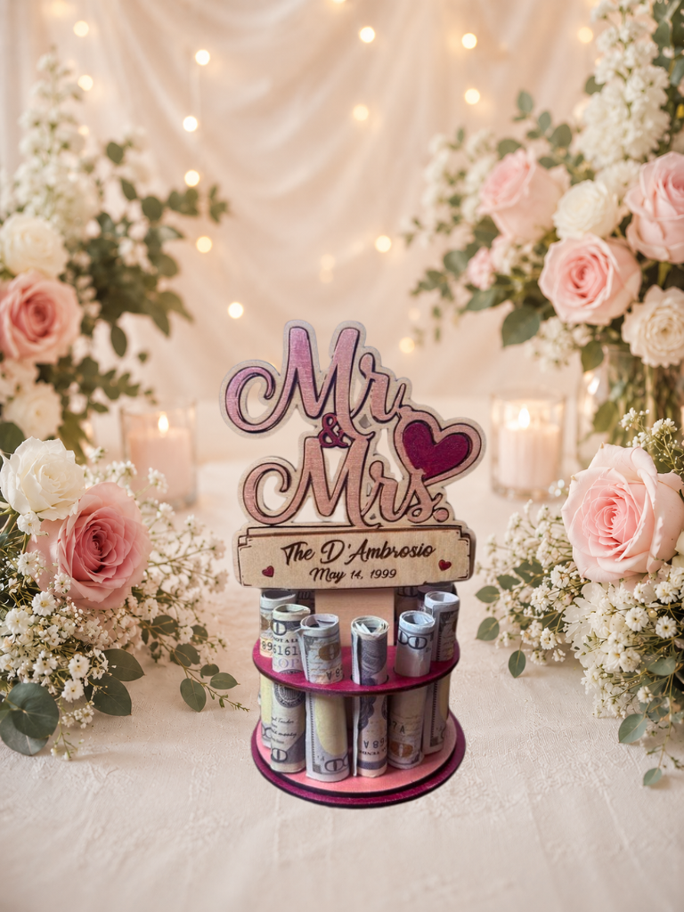 Premium handmade wood Mr. & Mrs. wedding money tree with laser-cut script lettering and personalized name banner reading "The D'Ambrosio May 14, 1999," displayed on a two-tiered pink circular base holding 12 rolled bills, surrounded by pink roses and candlelight