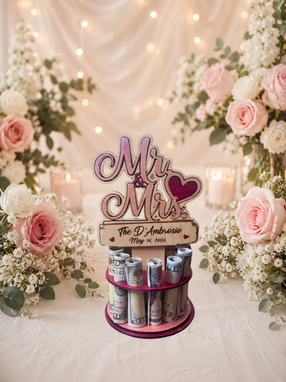 Premium handmade wood Mr. & Mrs. wedding money tree with laser-cut script lettering and personalized name banner reading "The D'Ambrosio May 14, 1999," displayed on a two-tiered pink circular base holding 12 rolled bills, surrounded by pink roses and candlelight