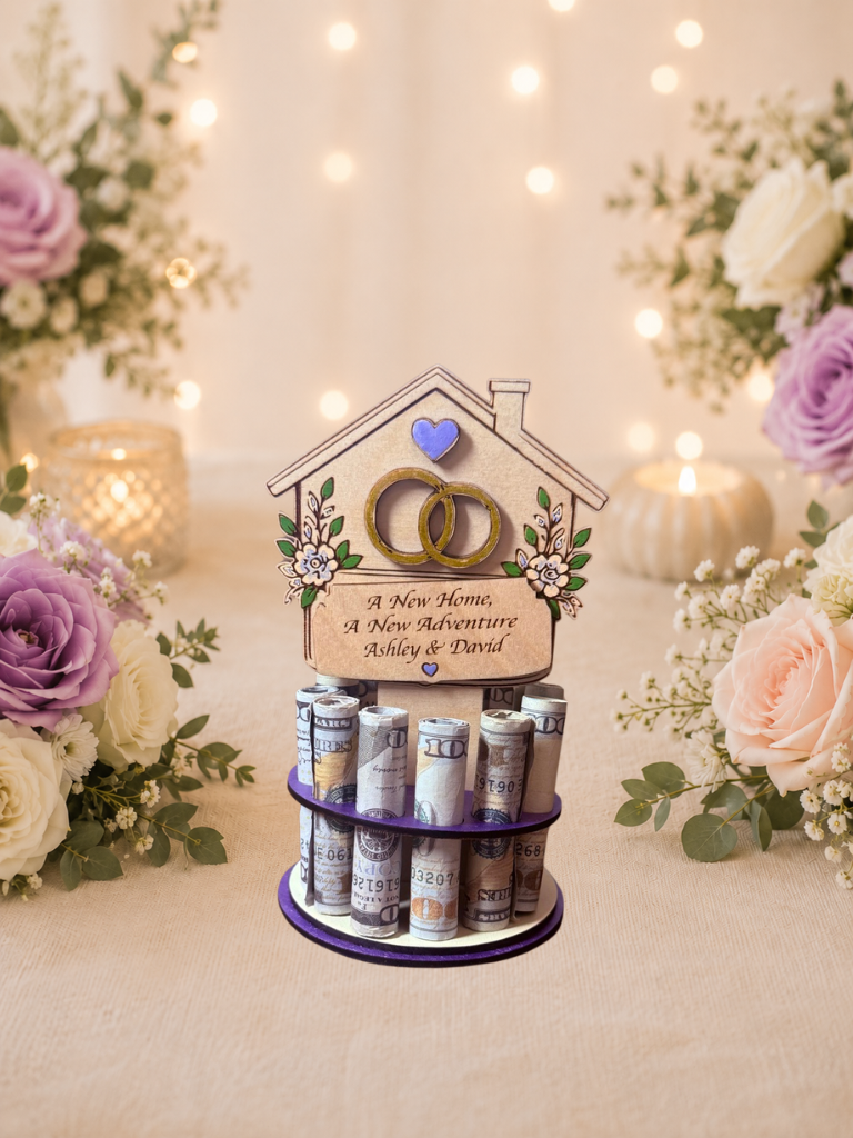 Premium handmade wood new home money tree with house-shaped topper featuring interlocking wedding rings, purple heart accent, and hand-painted florals with personalized text reading "A New Home, A New Adventure" and "Ashley & David," displayed on a single-tiered purple circular base holding 12 rolled bills, surrounded by purple and blush roses with candlelight