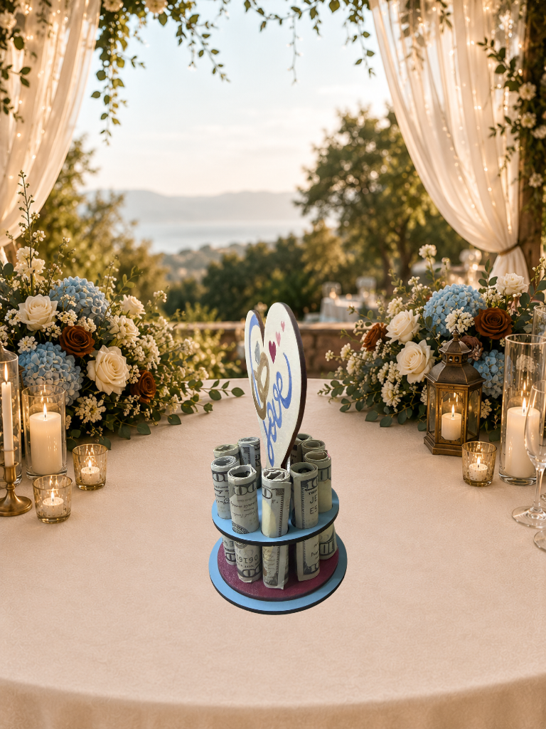 Handmade wood wedding money tree with a white heart-shaped top hand-painted with wedding rings and "Love" in blue script, set on a tiered circular base holding rolled bills, styled on an outdoor wedding gift table with blue hydrangeas and candlelight