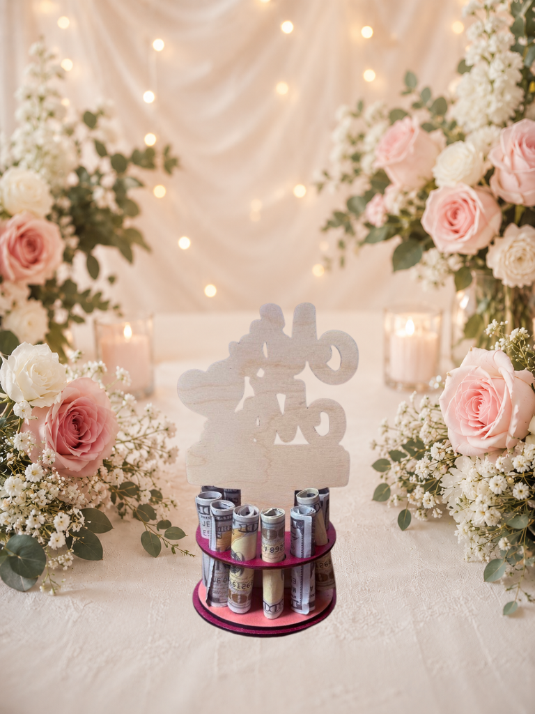 Premium handmade wood Mr. & Mrs. wedding money tree with laser-cut script lettering and personalized name banner reading "The D'Ambrosio May 14, 1999," displayed on a two-tiered pink circular base holding 12 rolled bills, surrounded by pink roses and candlelight
