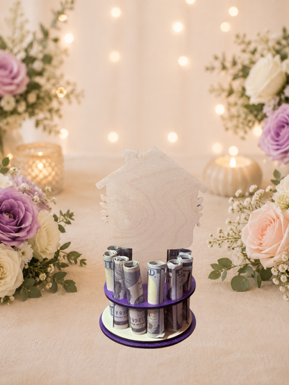 Premium handmade wood new home money tree with house-shaped topper featuring interlocking wedding rings, purple heart accent, and hand-painted florals with personalized text reading "A New Home, A New Adventure" and "Ashley & David," displayed on a single-tiered purple circular base holding 12 rolled bills, surrounded by purple and blush roses with candlelight