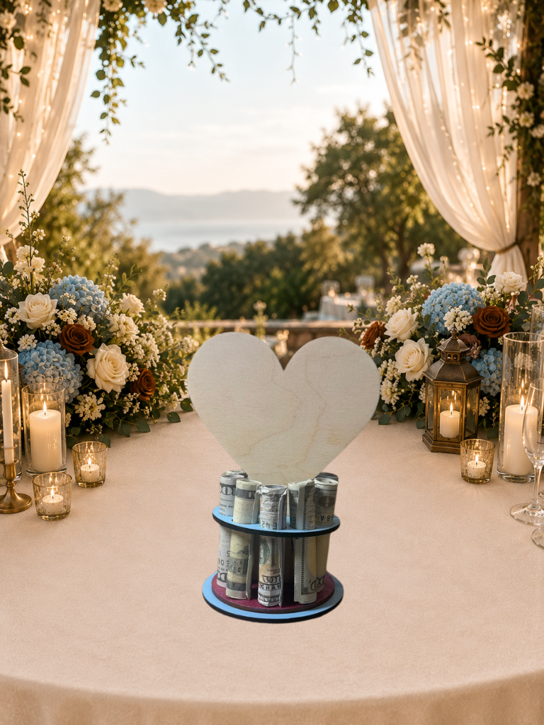 Handmade wood wedding money tree with a white heart-shaped top hand-painted with wedding rings and "Love" in blue script, set on a tiered circular base holding rolled bills, styled on an outdoor wedding gift table with blue hydrangeas and candlelight