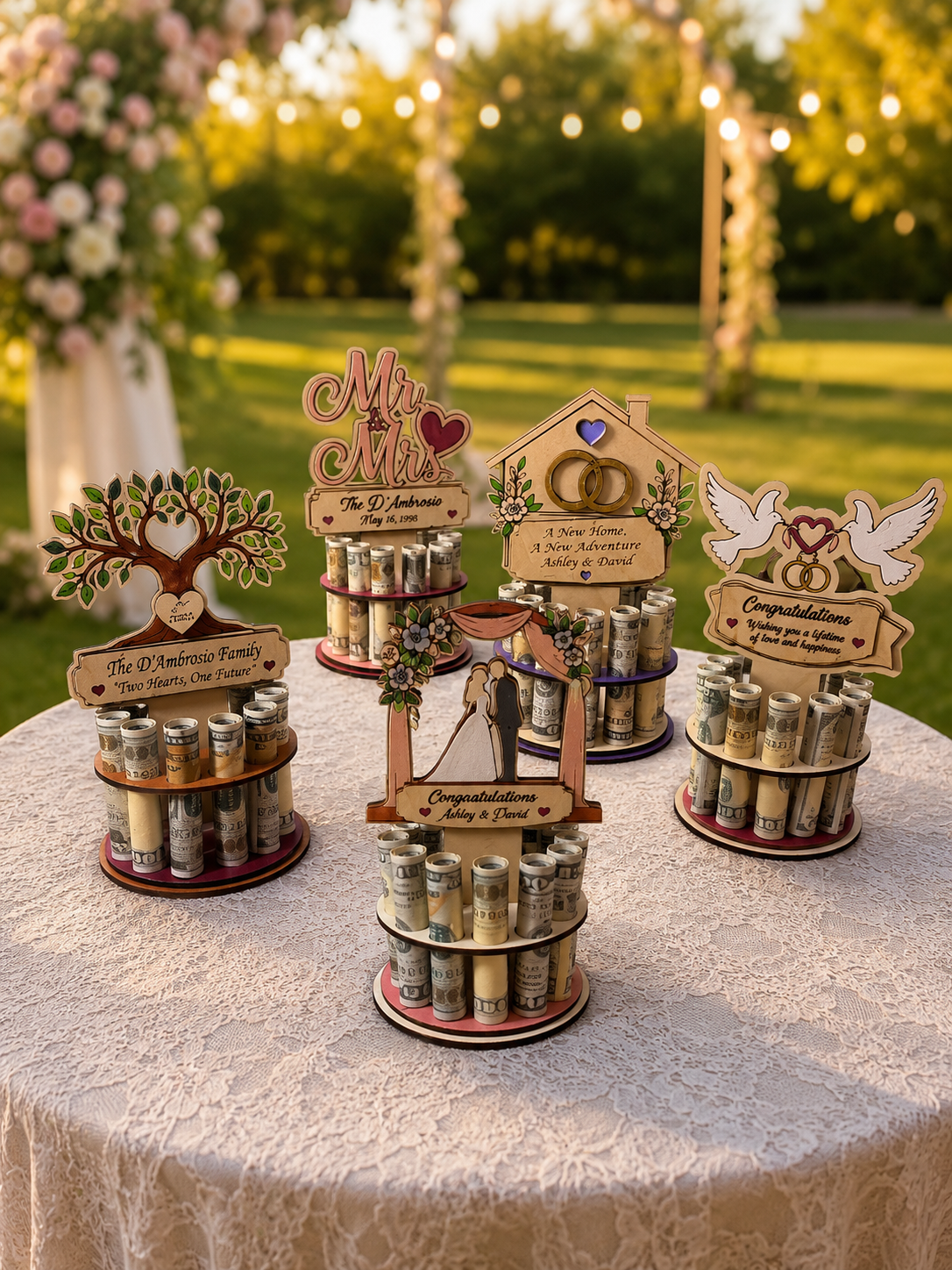 Five premium handmade wood wedding money trees displayed together on a lace-covered outdoor table — Tree of Life, Mr. & Mrs., New Home, Wedding Arch, and Dove designs — each holding 12 rolled bills, made from 1/4 inch Baltic birch, styled in a garden wedding setting