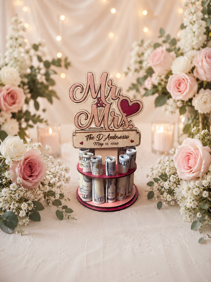 Premium handmade wood Mr. & Mrs. wedding money tree with laser-cut script lettering and personalized name banner reading "The D'Ambrosio May 14, 1999," displayed on a two-tiered pink circular base holding 12 rolled bills, surrounded by pink roses and candlelight