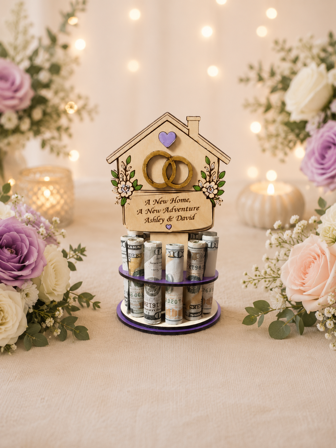 Premium handmade wood new home money tree with house-shaped topper featuring interlocking wedding rings, purple heart accent, and hand-painted florals with personalized text reading "A New Home, A New Adventure" and "Ashley & David," displayed on a single-tiered purple circular base holding 12 rolled bills, surrounded by purple and blush roses with candlelight