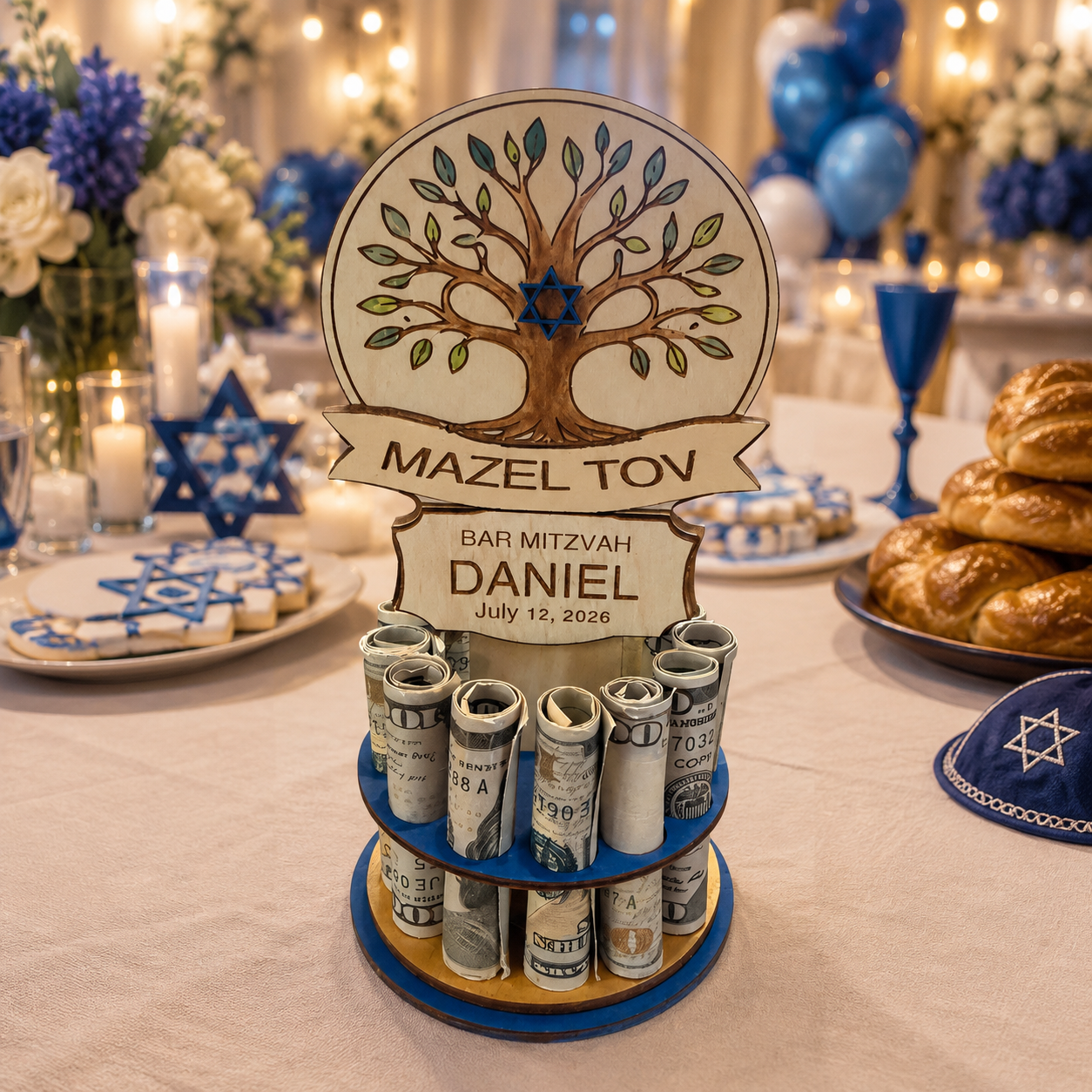Premium handmade wood tree of life Bar Mitzvah money tree with hand-painted tree, Star of David, Mazel Tov banner, and personalized name banner reading "BAR MITZVAH DANIEL July 12, 2026," on a blue single-tiered base holding 13 rolled bills, styled on a celebration table with blue and white flowers, candles, and challah bread