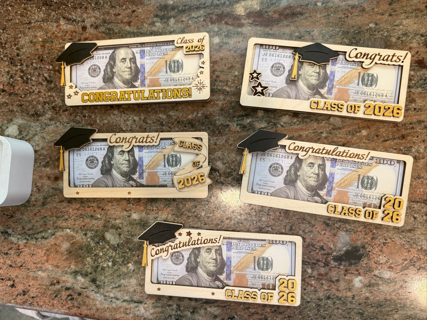 Five handmade layered Baltic birch wood graduation money holders displayed with bills, featuring a 3D graduation cap and gold Congratulations and Class of 2026 lettering in five different designs, approximately 6.5 to 7 inches long