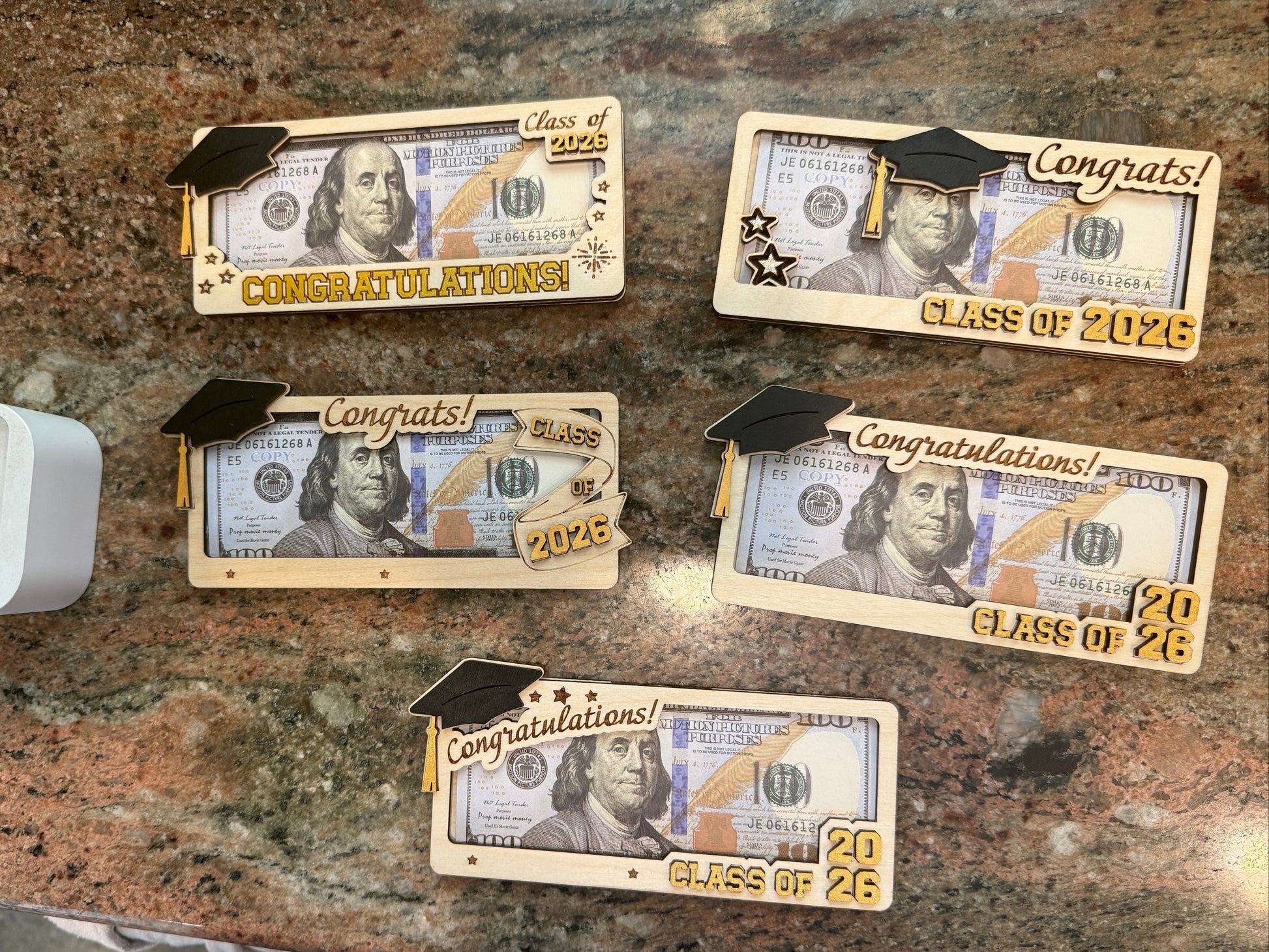 Five handmade layered Baltic birch wood graduation money holders displayed with bills, featuring a 3D graduation cap and gold Congratulations and Class of 2026 lettering in five different designs, approximately 6.5 to 7 inches long