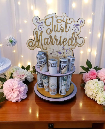 Handmade wood Just Married money tree with rolled hundred-dollar bills displayed in tiered rows, gold "Just Married" lettering on lavender tiers, styled with pink and white flowers and fairy lights