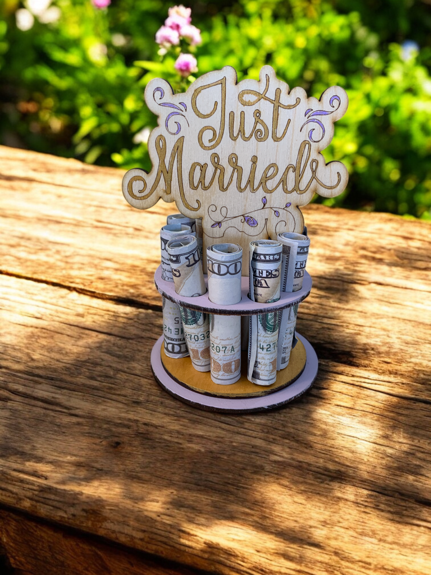 Handmade wood Just Married money tree with rolled hundred-dollar bills displayed in tiered rows, gold "Just Married" lettering on lavender tiers, styled with pink and white flowers and fairy lights