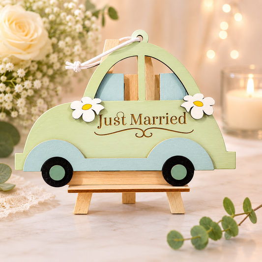 Handmade wood just married car gift card holder shown from the side in mint green and soft blue with white daisies and "Just Married" lettering, hanging from a white leather cord on a small wooden easel beside cream roses and eucalyptus