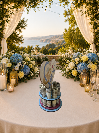 Handmade wood wedding money tree with a white heart-shaped top hand-painted with wedding rings and "Love" in blue script, set on a tiered circular base holding rolled bills, styled on an outdoor wedding gift table with blue hydrangeas and candlelight