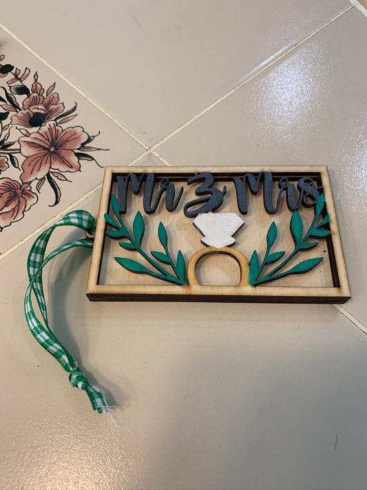 Handmade wood Mr & Mrs gift card holder with framed lettering, green laurel sprigs, and an engagement ring design, hanging from a green gingham ribbon on a tiled surface