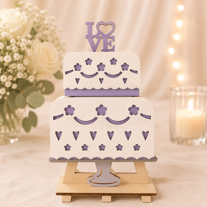 Handmade wood wedding cake gift card holder with two tiers, cut-out hearts and flowers in white and purple, topped with a "LOVE" cake topper, displayed on a small wooden easel beside white roses and a candle
