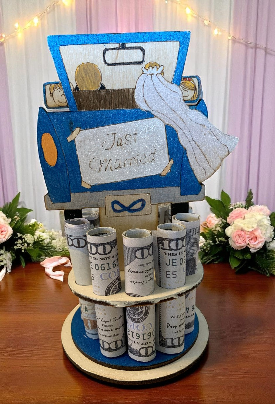 Handmade wood just married car money tree topped with a blue getaway car and bride's veil, displaying rolled hundred-dollar bills, styled with pink and white flowers and fairy lights