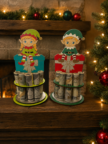 Two handmade wood Christmas elf money trees holding rolled bills, one with a girl elf on a teal gift box and one with a boy elf on a red gift box, hand-painted and displayed on a wooden mantel with a lit tree and garland