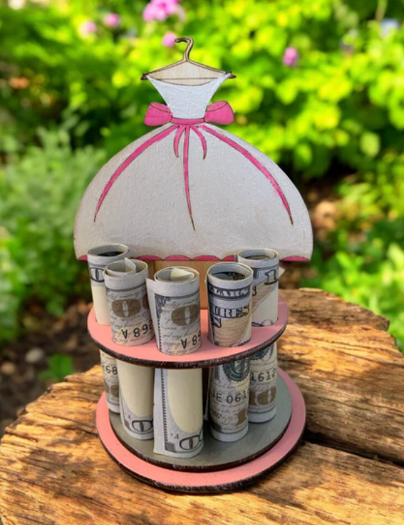 Handmade wood bridal shower money tree topped with a white bride's gown on a hanger with a pink bow, displaying rolled bills, photographed outdoors with greenery behind